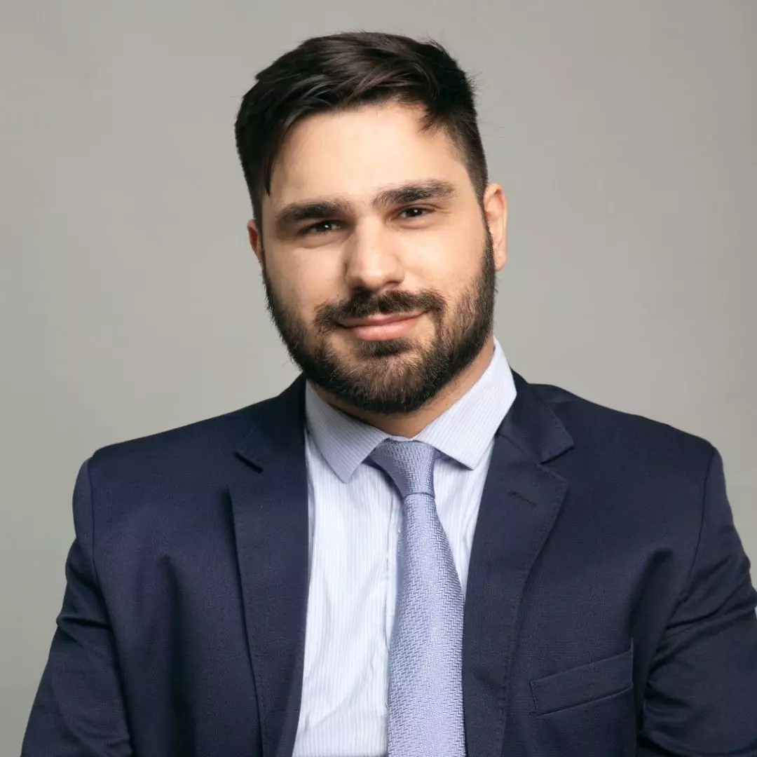 homem branco de cabelos castanhos curtos e usa barba. Ele veste blazer azul marinho, camisa branca e gravata cinza.