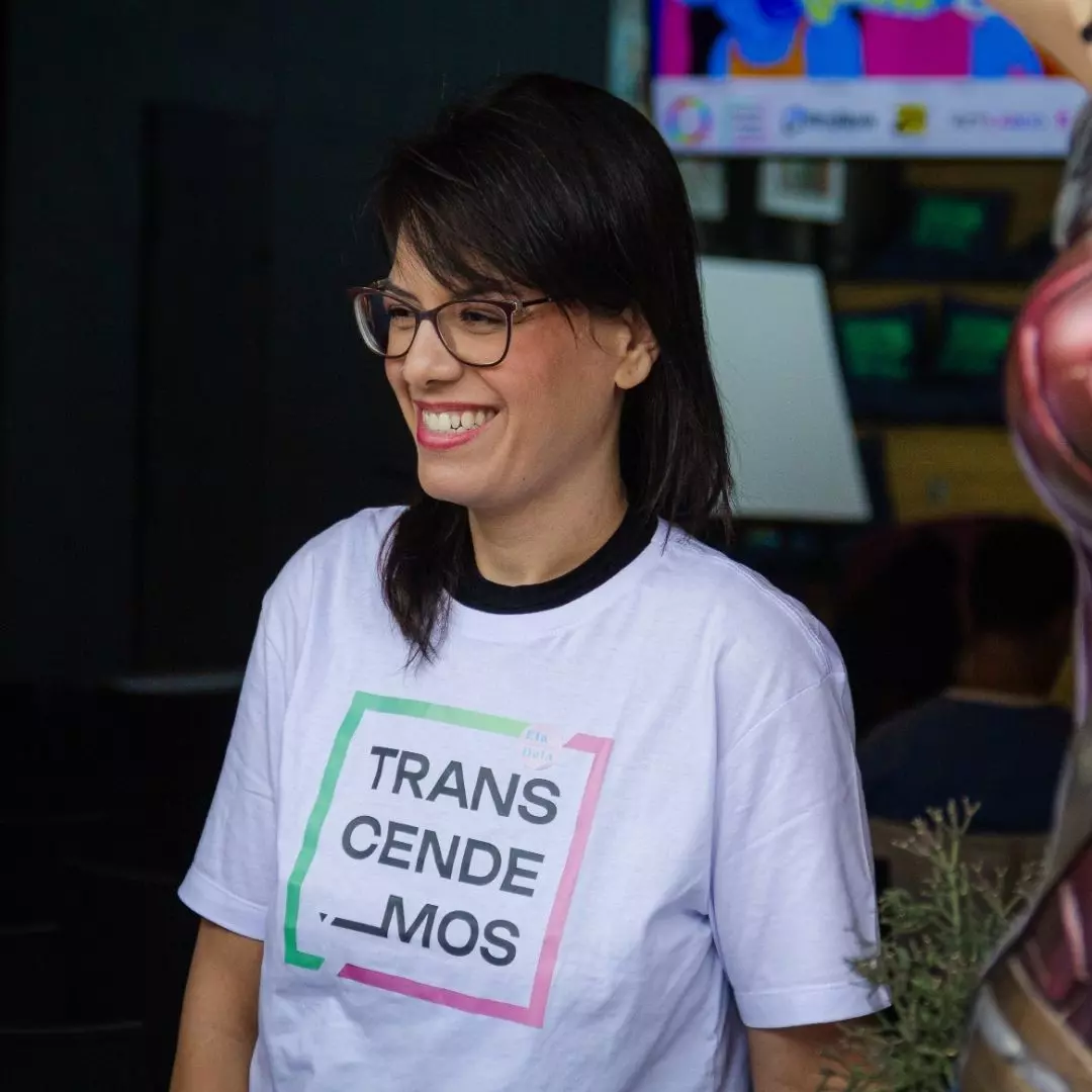 mulher branca de cabelos lisos castanhos. Ela usa óculos de grau e camiseta da Transcendemos e calça jeans.