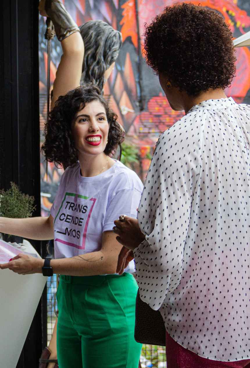 Simone Andrani em evento duas pessoas conversando. uma delas está de frente, é uma mulher branca com cabelos cacheados e camiseta da transcendemos e calça verde. A outra, de costas, está com camisa branca com bolinhas pretas.