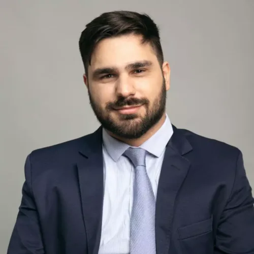 homem branco de cabelos castanhos curtos e usa barba. Ele veste blazer azul marinho, camisa branca e gravata cinza.
