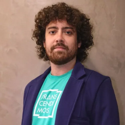 homem branco de cabelos cacheados castanhos e barba. Ele veste Blazer azul e camiseta verde da Transcendemos.