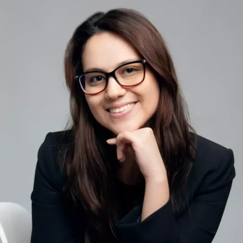 mulher branca, de cabelos castanhos lisos compridos, óculos de grau. Ela veste blazer preto e está com uma das mãos no queixo.