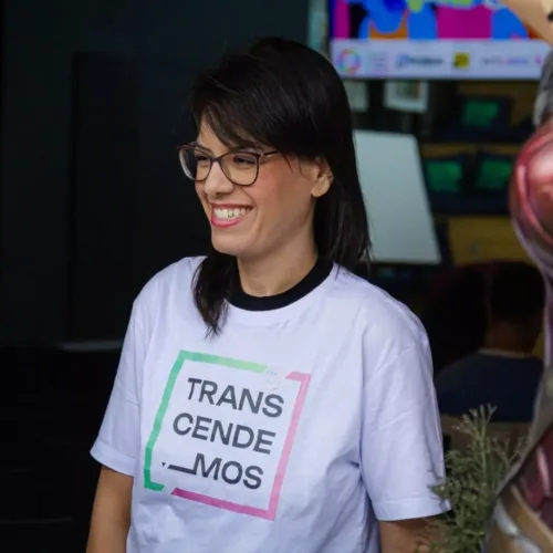 mulher branca de cabelos lisos castanhos. Ela usa óculos de grau e camiseta da Transcendemos e calça jeans.