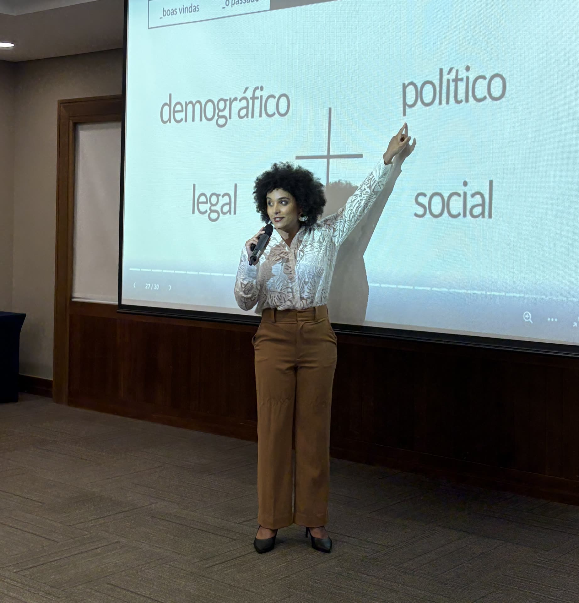 Gabriela Augusto em pé, com microfone, apresentando diante de uma tela de projeção com os termos demográfico, político, legal e social