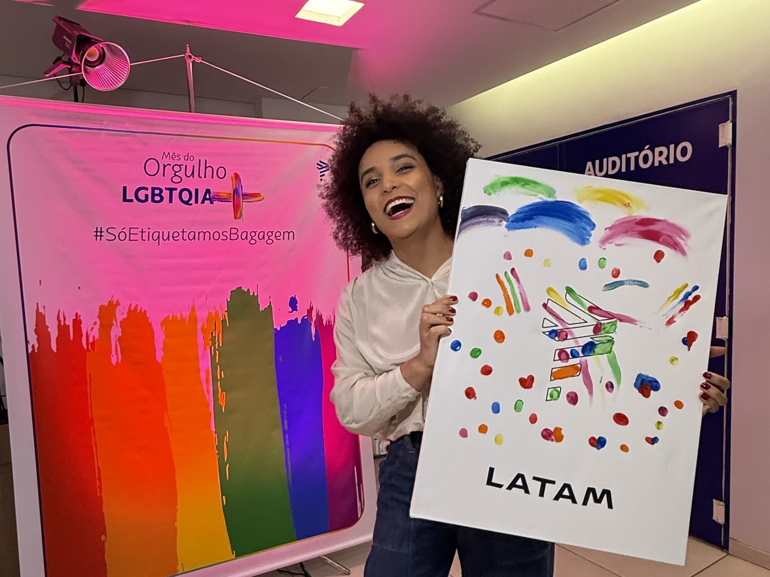 Gabriela Augusto sorrindo ao lado de um quadro pintado com o logo da LATAM, diante de painel do Mês do Orgulho LGBTQIA+