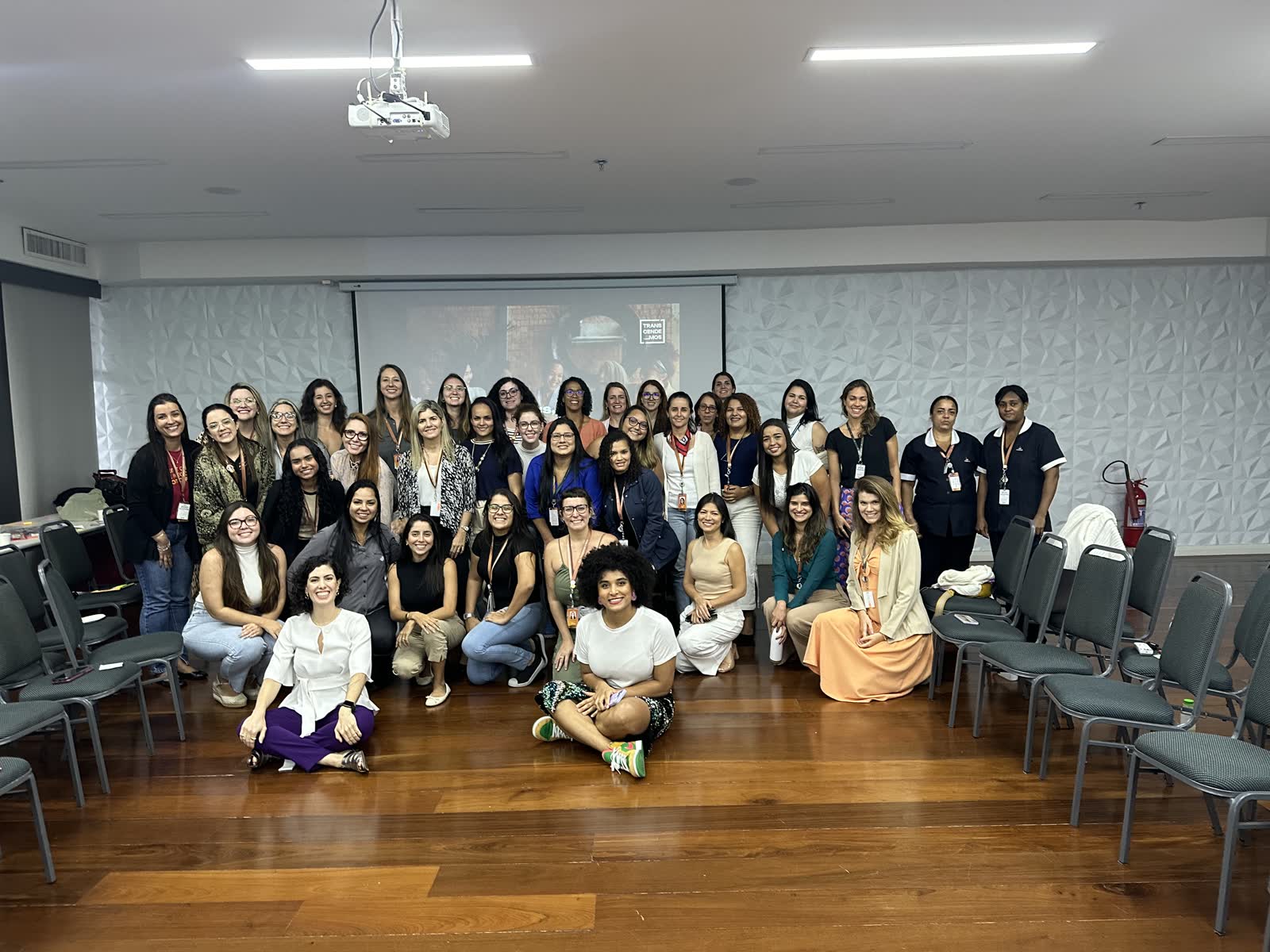 Grupo de colaboradoras da NTS reunido após o workshop Somos Múltiplas, posando para foto em frente a um telão com a marca da Transcendemos