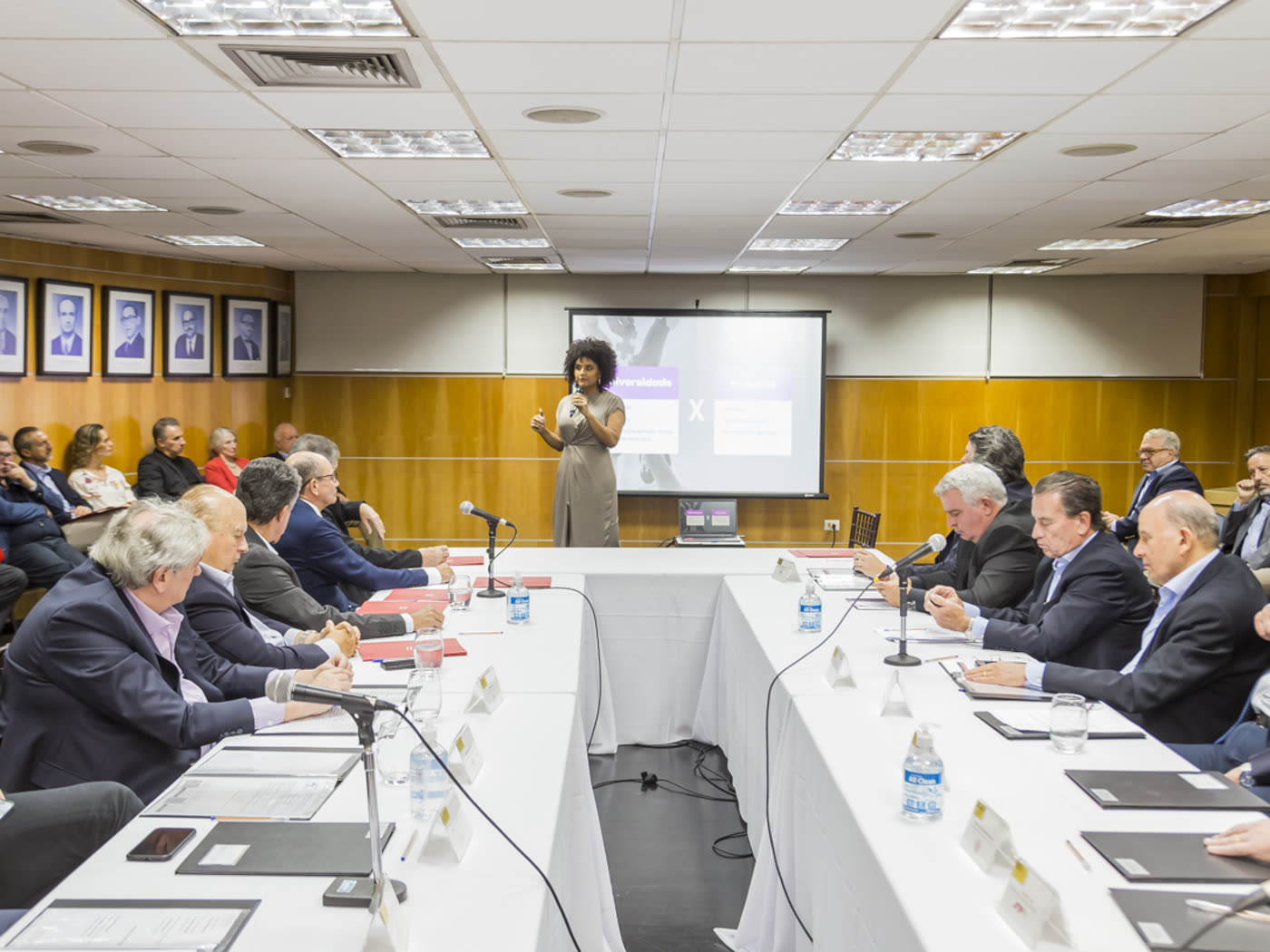 Gabriela Augusto conduz uma reunião de conselho em uma mesa composta quase inteiramente por homens de terno