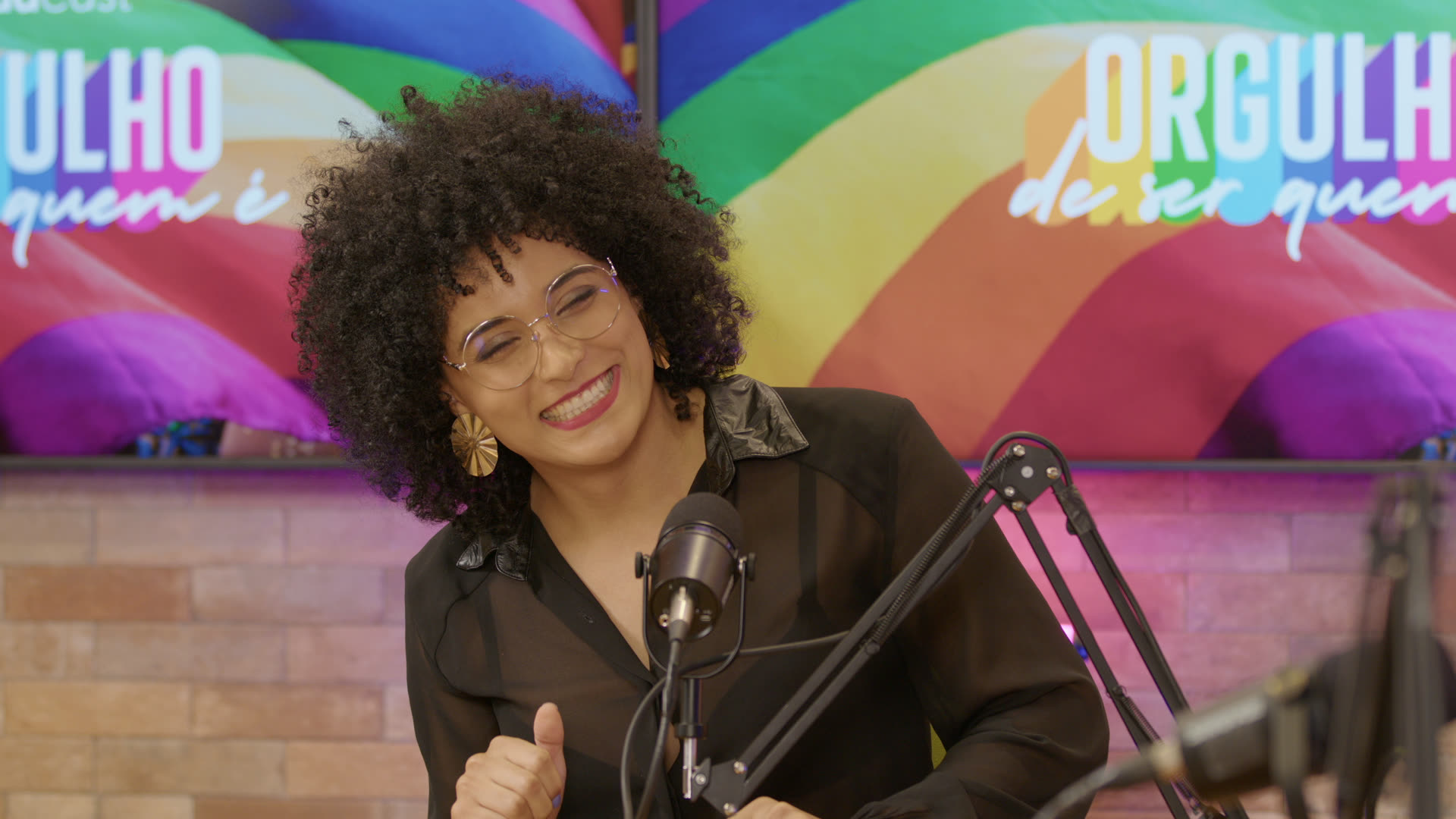 Gabriela Augusto sorrindo em transmissão ao vivo, diante de painel colorido com o texto Orgulho de ser quem é